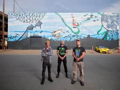 Three muralists stand proudly before their completed marine-themed artwork on a concrete wall in Southeast Portland's Central Eastside district. The vibrant mural depicts an underwater ecosystem with whales, fish, and maritime vessels rendered in blues and greens, while the artists - Jeremy Nichols in flannel and beanie, bigtroublehandpainted in black attire, and Devin Finley in work clothes - pose confidently on the industrial street. A yellow sports car and urban infrastructure frame the scene under an overcast Pacific Northwest sky.