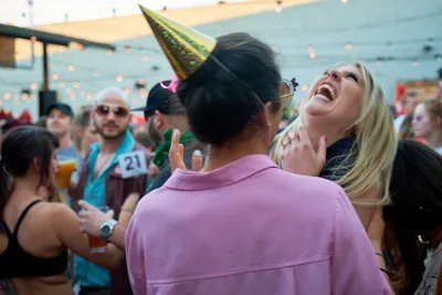 A blonde woman throws her head back in unrestrained laughter during Portland's Your Sunday Best day party at White Owl Social Club. The golden party hat catches the warm afternoon light as string lights create bokeh in the background. A man in a soft pink shirt leans in beside her, while fellow partygoers with drinks populate the festive outdoor scene.