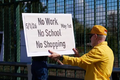 An activist in a vibrant yellow windbreaker and matching cap holds aloft a handwritten protest banner over Interstate 5 in North Portland's Skidmore neighborhood. The stark white sign bearing "5/1/26 No Work No School No Shopping May 1st" catches afternoon sunlight against the geometric shadows of a chain-link overpass fence. Industrial architecture frames this moment of grassroots organizing, as the demonstrator's weathered hands grip the edges of their call to action.