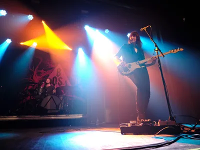 Emma Richardson commands the stage with her white bass guitar while Julian Dorio anchors the rhythm from behind his drum kit during Band of Skulls' 10th anniversary tour at Webster Hall in Manhattan. Dramatic stage lighting cuts through theatrical smoke, casting amber and cyan beams that illuminate the duo against the venue's shadowy backdrop. The intimate concert setting captures the raw energy of their milestone celebration, with Richardson positioned prominently in the foreground as colored spotlights create an atmospheric haze around the performance space.