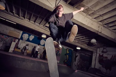 A skateboarder suspended mid-air executes a dynamic trick in the atmospheric basement of Commonwealth Skateboarding in Portland's Buckman district. The concrete space pulses with underground energy, its exposed wooden ceiling beams and colorful graffitied walls creating a raw, authentic backdrop. Dramatic lighting cuts through the subterranean setting, illuminating the athlete's focused expression and the worn grip tape of his deck as he floats momentarily above the industrial floor.