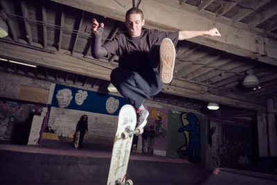 A skateboarder launches into a dynamic aerial maneuver inside Commonwealth Skateboarding in Portland's Buckman neighborhood. The raw concrete and exposed wooden beams of the indoor skate space frame the athlete mid-flight, while colorful skull artwork and graffiti murals create a gritty urban backdrop. The dramatic low-angle perspective captures the precise moment of weightless suspension, with the skater's weathered deck clearly visible beneath his sneakers.