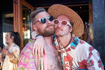 Two friends share an intimate moment at White Owl Social Club during Your Sunday Best's season opener in Portland, Oregon. The bearded man in reflective sunglasses embraces his companion, who sports white oval sunglasses and a woven straw hat alongside a vibrant floral Hawaiian shirt. Golden afternoon light filters through the venue's windows, creating a warm, celebratory atmosphere as other partygoers mingle in the background of this popular summer day party.