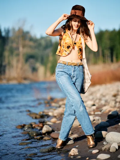 A young woman in bohemian attire strikes a carefree pose along the rocky riverbed of Oregon's Floodplain Trail in Multnomah County. She wears a vintage-inspired sunflower print vest over denim, her hands playfully adjusting a brown fedora as gentle waters flow past the pebbled shore behind her. The warm golden hour light bathes the scene, creating a dreamy atmosphere against the backdrop of distant evergreen forests and clear blue sky.