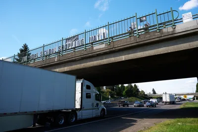 Activists from the Indivisible movement have suspended a protest banner reading "No Work School Shopping May 1st" across a concrete overpass spanning Interstate 5 in North Portland's Humboldt neighborhood. Below, a white semi-truck and steady stream of traffic flow beneath the weathered bridge structure, while afternoon sunlight filters through wispy clouds above. The grassroots demonstration transforms mundane infrastructure into a platform for labor organizing, capturing the intersection of highway commerce and social activism.
