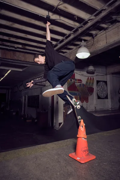 A skateboarder launches into a gravity-defying kickflip above a graffitied orange traffic cone in the industrial confines of Commonwealth Skateboarding in Portland's Buckman neighborhood. The raw concrete floor and exposed ceiling beams create a gritty backdrop, while weathered murals and pendant lighting cast dramatic shadows across this underground skating sanctuary. The athlete's black attire contrasts sharply against the weathered wooden walls, capturing a moment of pure athletic poetry in motion.