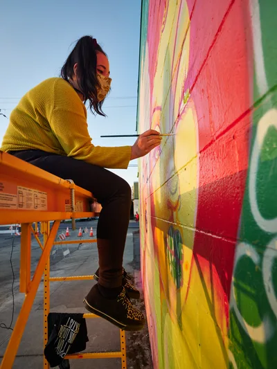 Multidisciplinary artist Vilatose works methodically on her mural 'Jardín Cubana' depicting three female figures, perched on an orange scissor lift against the expansive wall of a building in Portland's Alberta Arts District. Golden hour light bathes the scene as she adds delicate details to the rainbow-hued artwork featuring coral and yellow paint strokes, her leopard print face mask and mustard sweater creating a warm counterpoint to the vivid greens and pinks cascading across the concrete canvas. The industrial lift and scattered traffic cones below speak to the organized chaos of public art creation in this culturally rich Northeast Portland neighborhood.