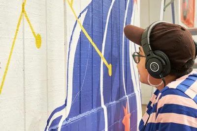 Mexican-American artist Maria Rodriguez, known as Sparkykneecap, focuses intently on her vibrant mural "Let's Talk" at Open Signal in Portland's Eliot neighborhood. Wearing Audio-Technica headphones and a navy striped shirt, she works on bold purple and yellow geometric forms painted on concrete walls. The commissioned piece explores themes of identity and culture while maintaining her signature playful aesthetic, bringing color and conversation to this Northeast Martin Luther King Jr. Boulevard arts venue.