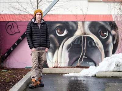 Artist Wetiko stands confidently before his striking mural of a pug's face at the Clay Creative Building in Southeast Portland's Hosford-Abernethy neighborhood. The dramatic close-up of the dog's expressive brown eyes and wrinkled snout dominates the pink wall behind him, creating a powerful juxtaposition with the artist's contemplative pose. Winter light filters through bare branches overhead, casting a subdued atmosphere over the scene while patches of snow dot the urban landscape.