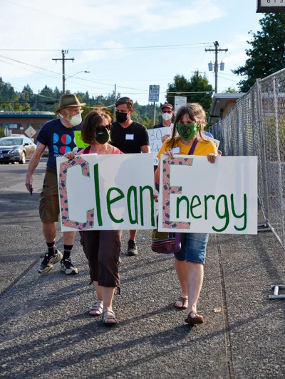 A diverse group of masked protesters marches along McLoughlin Promenade in Oregon City, carrying handmade banners spelling 'Clean Energy' in bold green lettering. The afternoon demonstration unfolds against a backdrop of power lines and suburban infrastructure, with participants ranging from an elderly bearded man in a fedora to young activists in casual summer attire. The scene captures the grassroots nature of environmental advocacy, as shadows stretch across weathered asphalt while demonstrators make their way toward Representative Kurt Schrader's office.