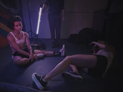 Professional skateboarder Christiana Means sits cross-legged on the concrete floor of Stronger Skatepark in Milwaukie, Oregon, her tattooed arms relaxed as she takes a break between sessions. The moody purple and magenta lighting from studio equipment casts dramatic shadows across the industrial space, while a photographer works nearby capturing the scene. The intimate moment captures the quiet intensity of skate culture, with Nike sneakers and scattered gear telling the story of dedicated practice.