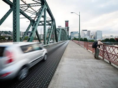 A solitary figure in a sun hat leans against the distinctive red pedestrian railing of Portland's iconic Hawthorne Bridge, gazing toward the city skyline as traffic streams past in a blur of motion. The weathered green steel truss structure frames the scene under overcast Pacific Northwest skies, while downtown Portland's modern towers rise beyond the Willamette River. The composition captures the contrast between human stillness and urban velocity, with the bridge's industrial architecture serving as a timeless connector between the city's east and west sides.