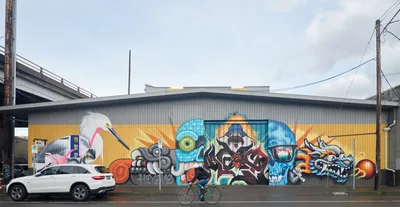 A vibrant street art mural by artist Heysus dominates the corrugated metal facade of an industrial warehouse on Water Avenue in Portland's Central Eastside. The expansive artwork features a kaleidoscope of characters including a white heron, blue skull, mythological figures, and abstract elements rendered in brilliant oranges, blues, and yellows against the building's golden lower wall. A cyclist pedals past the artwork while a white SUV is parked nearby, creating a dynamic urban scene beneath power lines and an overcast Pacific Northwest sky.