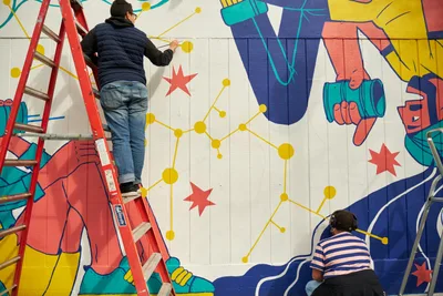 Two muralists work collaboratively on an expansive public art installation at Open Signal in Portland's Eliot neighborhood. One artist balances on a red ladder while adding delicate yellow constellation-like details to the vibrant composition, while another works at ground level on the lower portion of the wall. The mural features bold geometric forms in coral, turquoise, and navy blue, interwoven with red stars and connecting golden lines that create a sense of cosmic unity across the white concrete block surface.