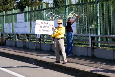 Two activists stand on a concrete pedestrian overpass above Interstate 5 in North Portland's Humboldt neighborhood, displaying a handwritten protest sign calling for a May 1st general strike. The man in a yellow shirt and baseball cap holds the white placard reading 'No Work No School No Shopping May 1st' while his companion in denim overalls raises his arm toward passing traffic below. Golden afternoon light casts long shadows across the bridge's weathered concrete surface, while green metal fencing and industrial structures frame this moment of grassroots political organizing. The urban landscape stretches behind them, punctuated by mature trees and the geometric patterns of highway infrastructure.