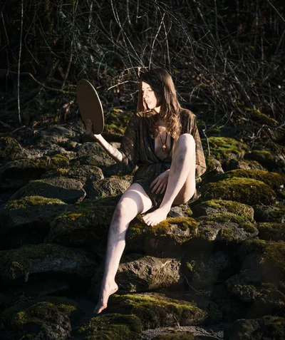 A young woman sits barefoot on moss-covered volcanic rocks along a riverbed on Oregon's Floodplain Trail, holding a circular mirror that catches the golden afternoon light. Her flowing brown hair and earth-toned dress harmonize with the ancient forest setting of bare winter branches and emerald moss. The warm sunlight creates dramatic shadows across the rocky terrain, while she gazes thoughtfully into the distance, creating an ethereal moment of self-reflection in nature.