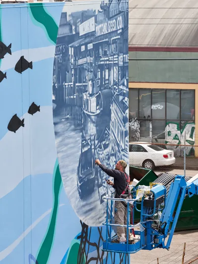 Jeremy Nichols and Devin Finley work from a blue hydraulic lift, adding intricate details to a large-scale monochromatic mural depicting historic streetscapes in Southeast Portland. The massive wall art transforms an urban corridor along Southeast 2nd Avenue in the Central Eastside district, where contemporary street art meets industrial architecture. Against a backdrop of chain-link fencing and parked vehicles, the artists methodically bring their vision to life under bright daylight, their collaborative effort representing the Portland Street Art Alliance's mission to enliven the city's built environment.