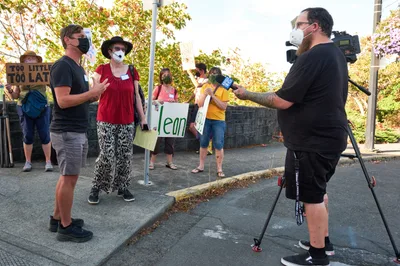 Michael Gaskill stands in animated conversation with a KOIN Channel 6 news crew on the sun-dappled sidewalk outside Representative Kurt Schrader's Oregon City office. Behind him, masked demonstrators hold handmade signs including "Too Little Too Late" and "Clean," their colorful clothing creating a vibrant backdrop against the autumn foliage. The golden afternoon light filters through leafy trees, casting warm shadows across the concrete as the interview unfolds during this climate change protest.