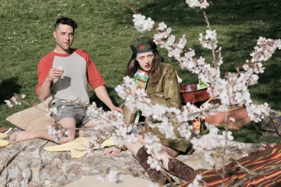 Two models pose for an editorial photography session beneath blooming cherry trees at Mount Tabor Park in Portland, Oregon. The woman, wearing a vintage military-style cap and golden jacket, sits alongside a man in a coral baseball tee as delicate pink and white blossoms frame the foreground. Soft afternoon light filters through the canopy, casting gentle shadows across their blanket spread with snacks and drinks on the verdant hillside.