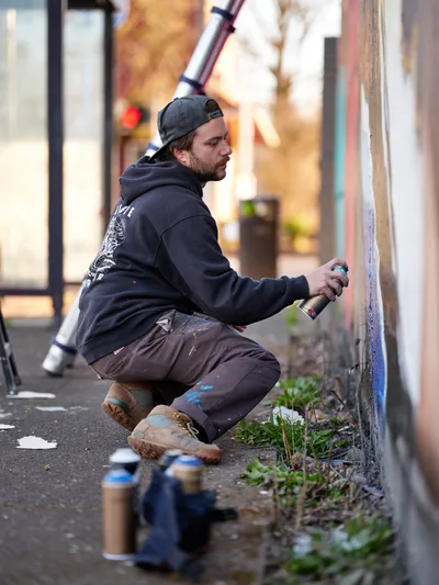Travis Fields, known as campographic, crouches in focused concentration as he works on a mural depicting poet Amanda Gorman at the intersection of Northeast 15th Avenue and Killingsworth Street in Portland's Alberta district. The artist, wearing a dark hoodie and backwards cap, grips a spray paint can while surrounded by an array of colorful aerosol cans scattered on the asphalt. Golden afternoon light filters through the urban streetscape, casting warm tones across the scene as Fields transforms the concrete wall into a tribute to the celebrated inaugural poet.