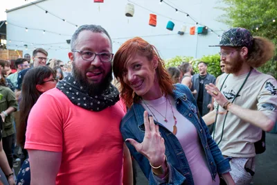 Two friends embrace the carefree spirit of Portland's summer day party scene under the dappled shade of White Owl Social Club's outdoor tent. The bearded man in coral pink and polka-dot bandana grins alongside a red-haired woman in denim who playfully flashes a peace sign, their joy infectious against the backdrop of fellow revelers. String lights overhead and colorful fabric bunting create an intimate festival atmosphere, while the crowd behind them pulses with the energy of Your Sunday Best's season opener.
