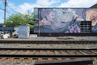 A vibrant street mural depicting an ethereal goddess figure adorned with intricate mandala headdress dominates the side of an industrial building in Portland's Buckman neighborhood. The artwork features flowing purple and coral-toned waves interwoven with ornate geometric patterns, creating a dreamlike narrative across the weathered concrete wall. Railway tracks cut through the foreground, their steel rails gleaming under the Pacific Northwest's filtered daylight, while power lines and urban infrastructure frame the scene. The juxtaposition of sacred feminine imagery against the industrial railway corridor captures the transformative power of public art in urban spaces.