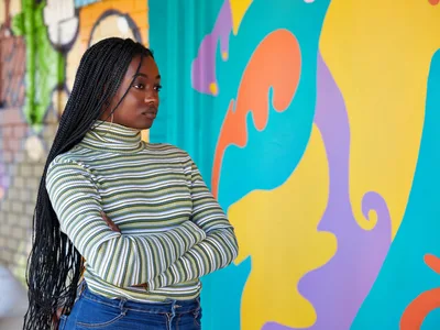 Naomi Likayi stands confidently before her completed mural in Portland's Alberta Arts District, her long braids cascading over a striped turtleneck as she poses with arms crossed. The dynamic street art behind her pulses with organic shapes in coral, turquoise, golden yellow, and violet, creating a bold backdrop that celebrates the neighborhood's creative renaissance. Soft daylight illuminates her contemplative expression, capturing a moment of artistic accomplishment against the kaleidoscopic wall that transforms the urban landscape.