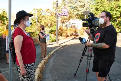 A masked climate activist in a red top and black hat speaks into a KOIN Channel 6 microphone during a rally outside Representative Kurt Schrader's Oregon City office. The bearded cameraman captures the interview against a backdrop of autumn foliage and suburban streets, while other masked demonstrators gather in the background. Golden afternoon light filters through the trees, creating a warm glow over this moment of grassroots political engagement.