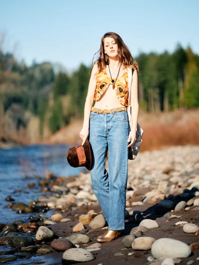 A young woman with flowing brown hair stands contemplatively along the rocky river bed of the Columbia River in Multnomah County, Oregon. She wears a distinctive animal-print crop top paired with high-waisted denim and carries a brown leather bag, her eyes gently closed as golden hour light illuminates her face. The river's gentle waters lap against smooth river stones while evergreen forests create a soft backdrop under the clear Pacific Northwest sky. Her bohemian styling and peaceful demeanor capture a moment of serene connection with Oregon's natural landscape.