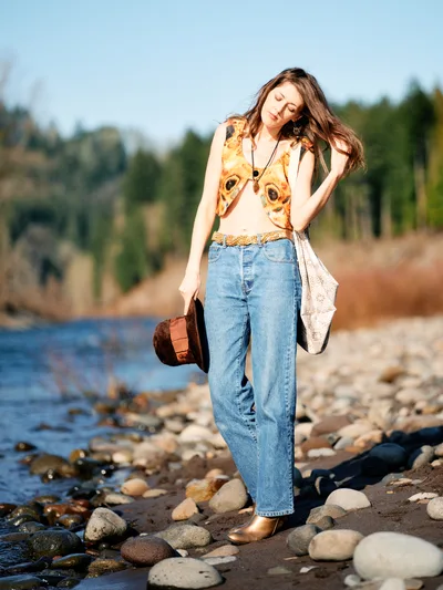 A young woman with flowing brown hair stands contemplatively along the rocky shoreline of the Willamette River in Oregon's Floodplain Trail. Bathed in warm golden hour light, she wears a distinctive graphic tank top featuring dog portraits, high-waisted denim, and carries a vintage brown leather satchel. Her peaceful expression and relaxed posture against the backdrop of smooth river stones and gentle waters create an intimate portrait of quiet reflection in nature.