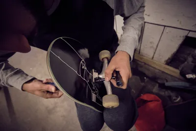 In the intimate confines of Commonwealth Skateboarding on Southeast Portland's 20th Avenue, hands work methodically over a black skateboard deck, adjusting trucks with practiced precision. The scene unfolds in warm, muted light that casts gentle shadows across weathered concrete floors, while red and gray hoodies frame the focused maintenance ritual. This quiet moment captures the behind-the-scenes dedication that keeps Portland's skate culture rolling.