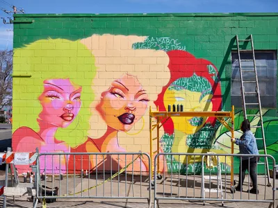 Multidisciplinary artist Vilatose works on 'Jardín Cubana,' a striking mural featuring three stylized female figures with vivid pink, yellow, and red hair against an emerald green cinderblock wall. The scene captures the creative energy of Portland's Alberta Arts District, with construction barriers and scaffolding framing the artist at work under clear blue skies. The mural showcases bold feminine portraits rendered in contemporary street art style, transforming an urban wall into a canvas of color and expression.