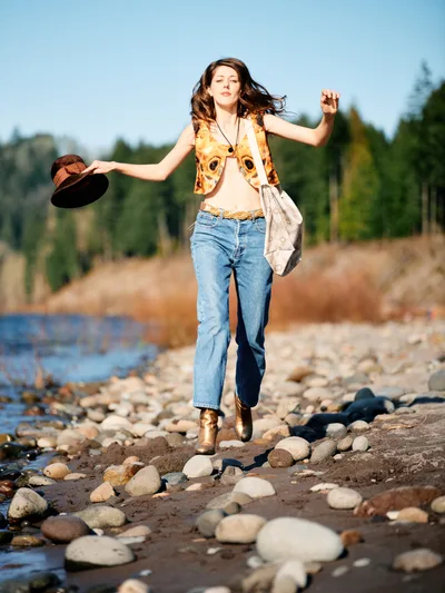 A young woman runs joyfully along the rocky shoreline of an Oregon waterway, her chestnut hair flowing in the golden evening light. She wears a vintage-inspired sunflower print vest over a white tank top, paired with classic blue jeans and leather boots, while swinging a brown fedora hat in one hand. The Floodplain Trail setting creates a dreamy backdrop with its smooth river stones, gentle water reflections, and towering Pacific Northwest forest.