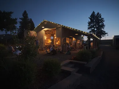 The exterior of the barn at Abbey Road Farm in Carlton, Oregon, photographed at dusk. The corrugated metal barn is strung with warm globe lights along its roofline, glowing invitingly against a deep blue twilight sky. Groups of guests relax on Adirondack chairs and benches on the gravel patio, socializing in the warm light spilling from the open barn interior. Tall evergreen trees flank the building, and lush garden plantings border a stone pathway in the foreground.