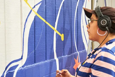 Maria Rodriguez, known as Sparkykneecap, works intently on a vibrant blue and yellow mural at Open Signal in Portland's Eliot neighborhood. The Mexican-American artist wears black headphones and a striped shirt while painting geometric shapes on the weathered wooden wall. Warm afternoon light illuminates the creative process as Rodriguez adds precise brushstrokes to the commissioned piece titled 'Let's Talk,' exploring themes of identity and culture through bold color and form.
