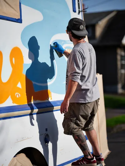 A muralist in a black baseball cap and gray long-sleeve shirt carefully applies vibrant blue paint to a food truck's exterior on Northeast 9th Avenue in Portland's King neighborhood. His tattooed leg and casual cargo shorts speak to the artistic street culture as he works under bright daylight, transforming the white vehicle with bold orange and blue graphics. The urban setting provides an authentic backdrop for this moment of creative commerce.