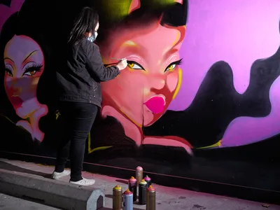 Multidisciplinary artist Vilatose meticulously applies detail to a striking mural featuring bold feminine faces rendered in electric pinks, purples, and yellows. Working in the intimate confines of Clay Creative's parking garage in Portland's Hosford-Abernethy neighborhood, the masked artist moves with practiced precision, surrounded by an arsenal of spray paint cans scattered across the concrete floor. The dramatic lighting illuminates both the artist's focused silhouette and the luminous portrait emerging on the wall, creating a compelling study of creative process in urban space.