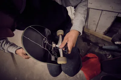 A skateboarder's weathered hands carefully adjust the trucks on a well-worn deck at Commonwealth Skateboarding in Portland's Buckman neighborhood. The moody lighting and intimate framing capture the ritual of board maintenance, with the skater's gray hoodie and striped sleeves creating visual texture against the dark skateboard. Red accents from clothing peek into the frame, adding warmth to the industrial setting of this Southeast Portland skate shop.