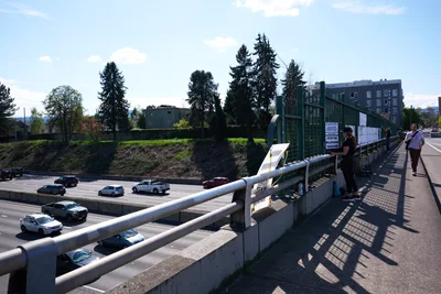Beneath a clear Pacific Northwest sky dotted with white clouds, activists from the Indivisible movement unfurl a protest banner from a pedestrian overpass spanning Interstate 5 in North Portland. The concrete bridge frames a scene of urban activism, with participants silhouetted against the bright afternoon light as traffic flows steadily below. Towering evergreens and modern residential buildings create a distinctly Portland backdrop, while the activists' coordinated banner drop transforms this everyday infrastructure into a stage for political expression.