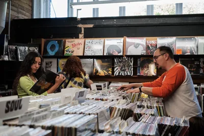 Three music enthusiasts browse through extensive vinyl collections at Music Millennium during the annual Record Store Day celebration in Portland's Kerns neighborhood. The woman in lime green examines album artwork while her curly-haired companion and a bearded man in orange systematically flip through alphabetized crates. Natural light filters through large windows, illuminating the store's carefully curated displays of classic and contemporary albums that line the walls above the browsing bins.