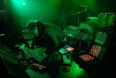 Bathed in supernatural emerald light, electronic musician Eli Savage hunches over his intricate array of synthesizers, drum machines, and glowing grid controllers during his ellipsism performance at RXFest 2019. The atmospheric green wash transforms Dante's intimate venue into a cybernetic cathedral, where analog knobs and digital displays pulse with multicolored frequencies. Savage's concentrated posture suggests deep communion with his electronic arsenal, while the moody lighting creates dramatic shadows that blur the line between performer and machine.