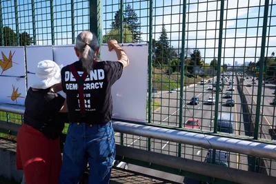 Two activists from the Indivisible movement position themselves on the North Skidmore Street overpass above Interstate 5 in Portland's Humboldt neighborhood, securing protest banners to the chain-link fence. The silver-haired man in a "Defend Democracy" t-shirt and his companion work methodically in the afternoon light, their white banner visible against the backdrop of moving traffic below. Evergreen trees frame the scene while cars flow steadily beneath this quiet act of civil engagement.