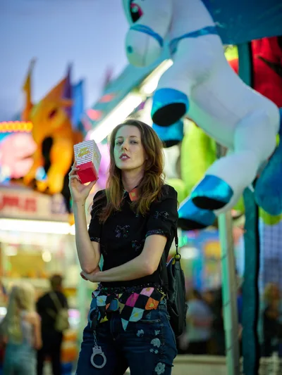 A young woman holds a red drink carton while standing beneath towering inflatable carnival decorations at the Clark County Fair in Vancouver, Washington. The evening scene pulses with electric blues, greens, and magentas from the midway's neon glow, creating a cinematic backdrop of childhood nostalgia and summer revelry. Her contemplative expression contrasts with the exuberant carnival atmosphere swirling behind her in soft focus.