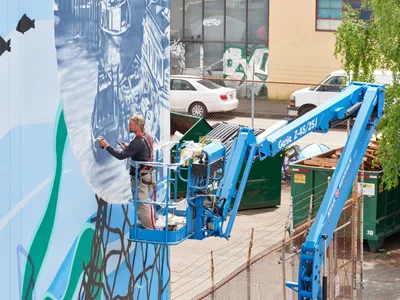 Jeremy Nichols and Devin Finley work methodically from a bright blue Genie aerial lift, applying paint to a sweeping mural on a building wall in Portland's Central Eastside district. The scene captures the industrial rhythm of street art creation, with construction debris and chain-link fencing framing the artists against their towering canvas of blues and greens. Afternoon light illuminates the working duo as they transform urban architecture into public art.