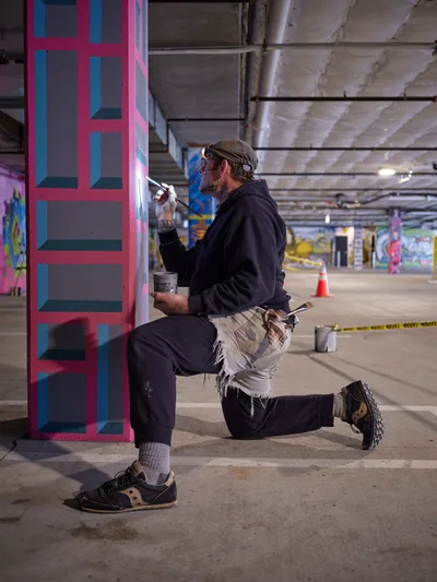 Portland artist and bike activist Rabbitt kneels in focused concentration as he paints his signature geometric pattern on a concrete column in the Clay Creative building's parking garage. The warm artificial lighting illuminates his work-worn hands applying pink and blue paint to create bold rectangular shapes, while the industrial space around him buzzes with creative energy. Paint cans and brushes scattered at his feet speak to the methodical process of transforming this utilitarian space into an artistic statement.