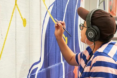 Maria Rodriguez, known as Sparkykneecap, applies vibrant blue and yellow paint to a wall at Open Signal in Portland's Eliot neighborhood. The Mexican-American artist wears headphones and a striped shirt while working on the commissioned mural titled "Let's Talk," their brush carefully adding flowing lines to the geometric composition. Warm afternoon light illuminates the creative process as Rodriguez transforms the white wall into a playful exploration of identity and community connection.