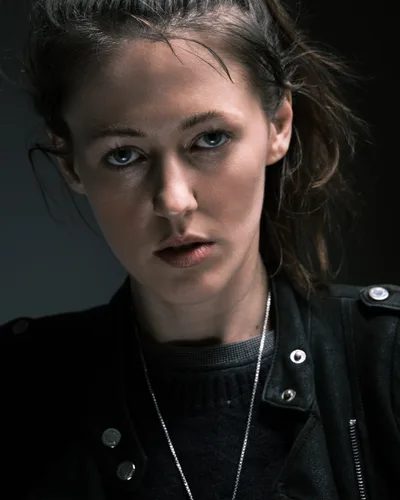 Rhianna Atwood gazes directly at the camera with piercing blue eyes in this intimate portrait captured in Portland's historic Irvington district. Tousled brown hair frames her face while she wears a black leather jacket over a dark mesh top, accented by a delicate silver chain necklace. The moody lighting creates dramatic shadows that emphasize the contemplative intensity of her expression against the neutral background.