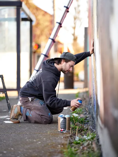 Travis Fields, known as campographic, crouches intensely beside a concrete wall on Northeast Killingsworth Street, spray paint can in hand as he works on a mural tribute to poet Amanda Gorman. The artist, dressed in paint-splattered clothing and a baseball cap, is captured mid-creation in the warm afternoon light of Portland's Alberta district. Scattered spray cans and small plants emerging from sidewalk cracks frame the intimate scene of artistic dedication.