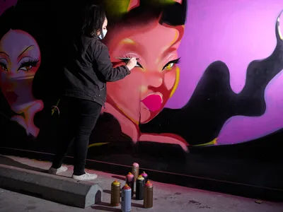 Multidisciplinary artist Vilatose meticulously details a striking magenta-toned portrait on the concrete wall of Clay Creative's parking garage in Portland's Hosford-Abernethy neighborhood. The masked artist, dressed in black, adds precise brushstrokes to luminous feminine faces that emerge from swirling purple and pink gradients. Scattered spray paint cans at her feet reflect the urban canvas transformed into gallery space, where raw industrial architecture meets contemporary street art.