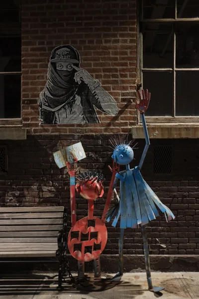 A striking urban tableau unfolds on Berry Street in Williamsburg, Brooklyn, where weathered brick walls frame a powerful dialogue between street art and sculptural installation. A monochromatic mural depicts a masked figure in contemplative pose, their piercing gaze watching over brightly painted metal sculptures below—a crimson jack-o'-lantern and azure figure with upraised arm create a vivid foreground against the industrial backdrop. Harsh daylight casts sharp shadows across the weathered sidewalk, emphasizing the raw texture of urban decay and creative resilience.